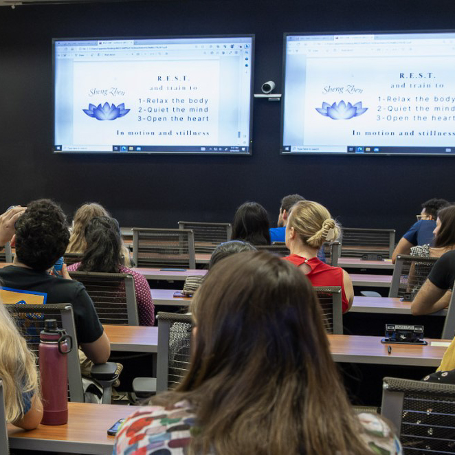 A group of people attending a presentation on relaxation techniques, with text on two screens displaying the principles of R.E.S.T. and a calming lotus flower.